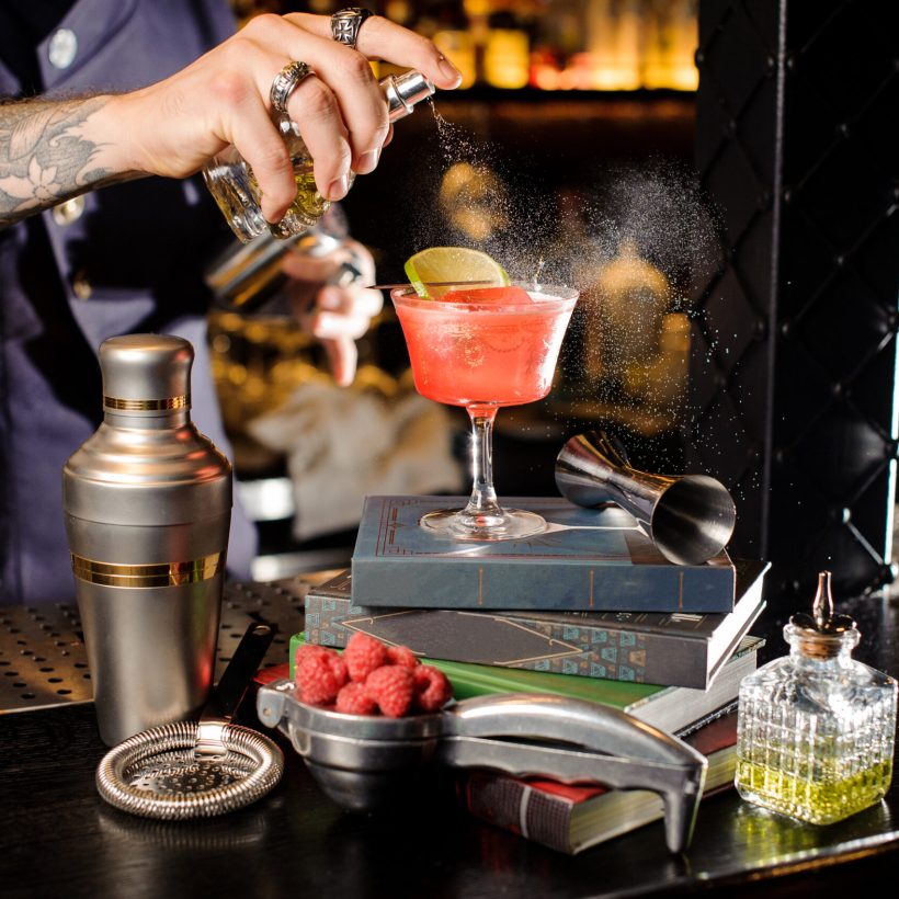 Barman spraying peat bitter on a glass with red sweet summer cocktail arranged on books on the bar counter